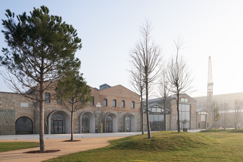 Brick facade with arched openings facing a gravel plaza lined with bare winter trees