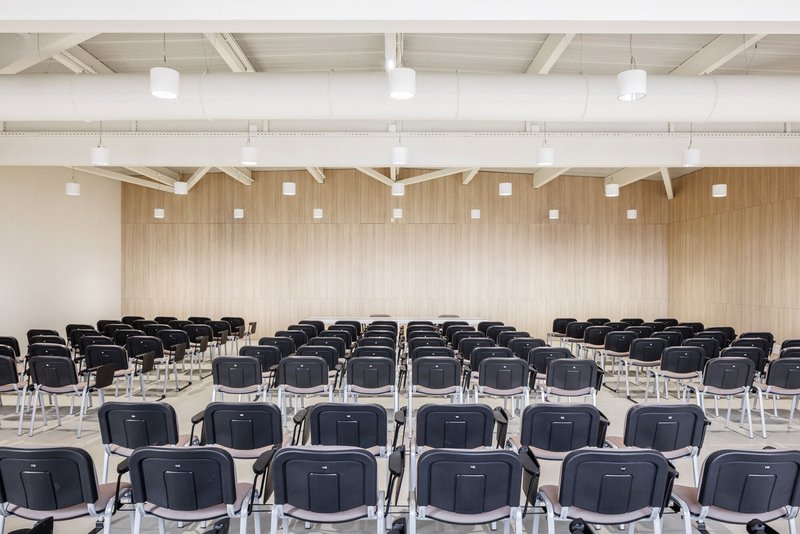 Assembly hall with rows of black chairs facing a vertical timber-clad wall under exposed white trusses
