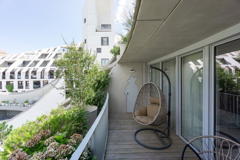 Timber deck balcony with hanging chair, planter bed, and concrete soffit overhead in daylight