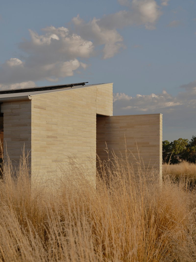 Two masonry volumes in contrasting tones emerging from dry grasses under a cloudy sky