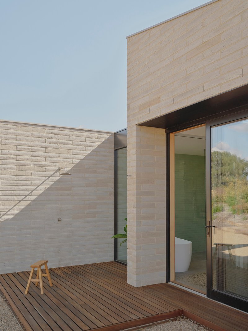 Timber deck courtyard enclosed by cream brick walls with sliding glass door to bathroom under bright sky