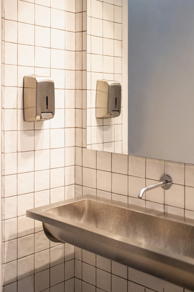 Corner washroom detail with white square tile and wall-mounted soap dispensers above a limestone trough sink
