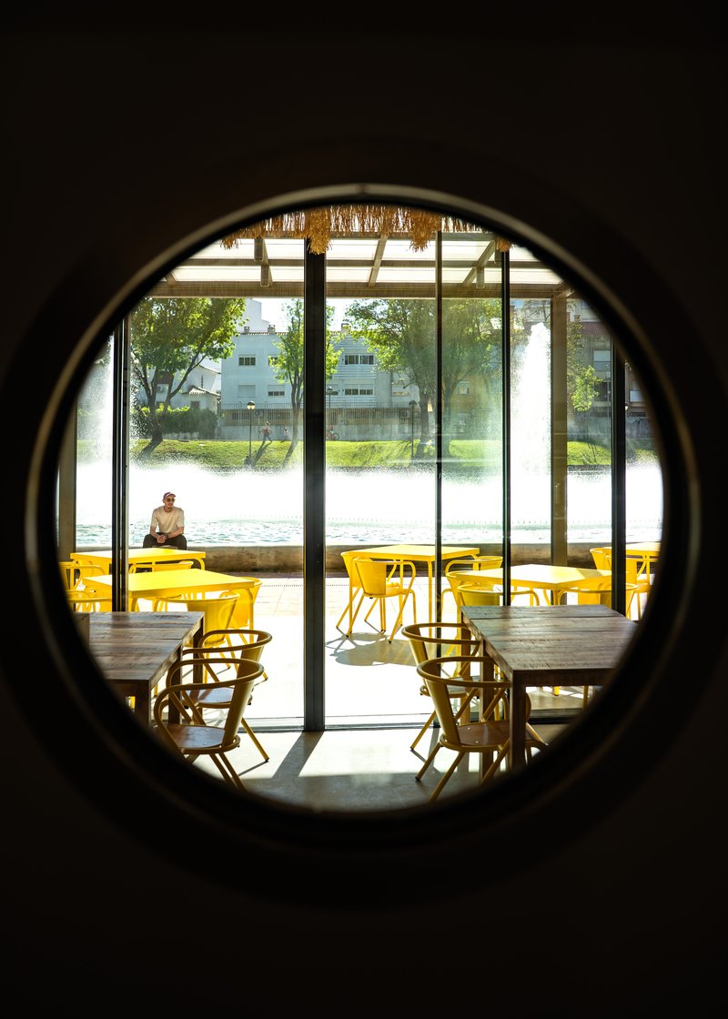View through circular portal opening to dining area with yellow seating and glazed facade beyond