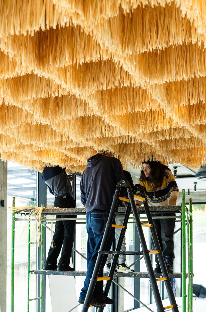 Workers on scaffolding installing layers of hanging straw or raffia panels beneath a ceiling structure
