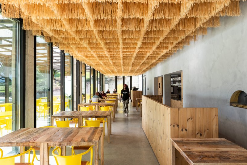 Dining hall with timber tables, yellow chairs, and hanging woven ceiling installation over a polished concrete floor