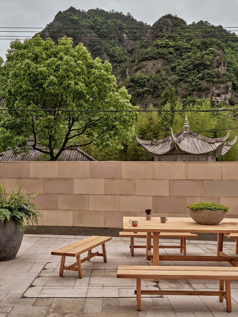Timber dining table and benches on paved terrace against sandstone wall with hillside beyond