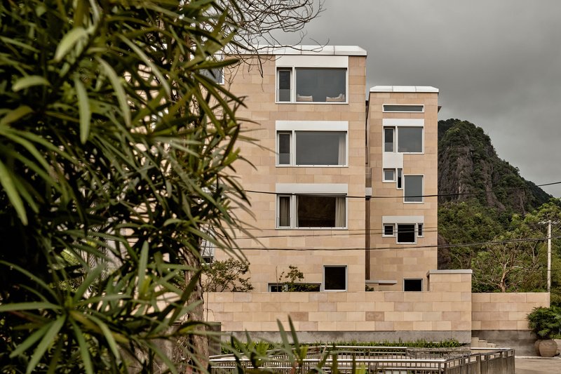Facade of stacked block volumes with punched windows framed by foreground foliage and a volcanic peak behind