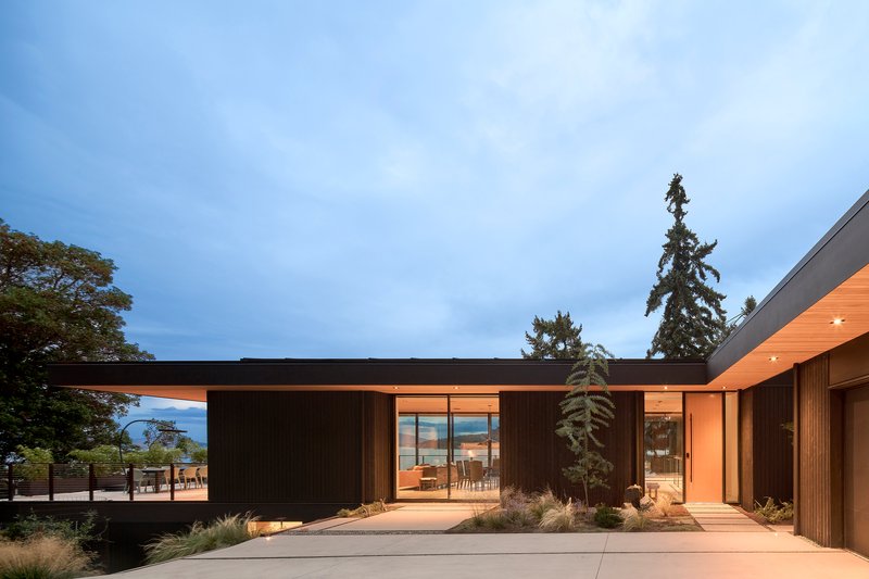 Courtyard entry with dark vertical cladding, timber soffit, and planted beds at dusk