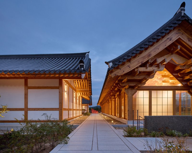 Concrete pathway between two tiled roof volumes with planted beds at twilight