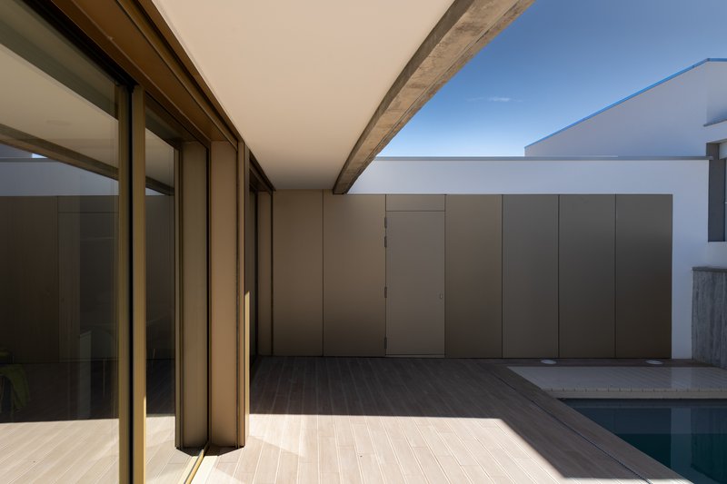 Covered terrace with sliding bronze-framed glass doors, timber decking and tan panel wall beside a pool
