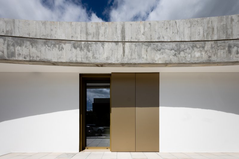 Curved white rendered wall with bronze-paneled door beneath a board-formed concrete parapet