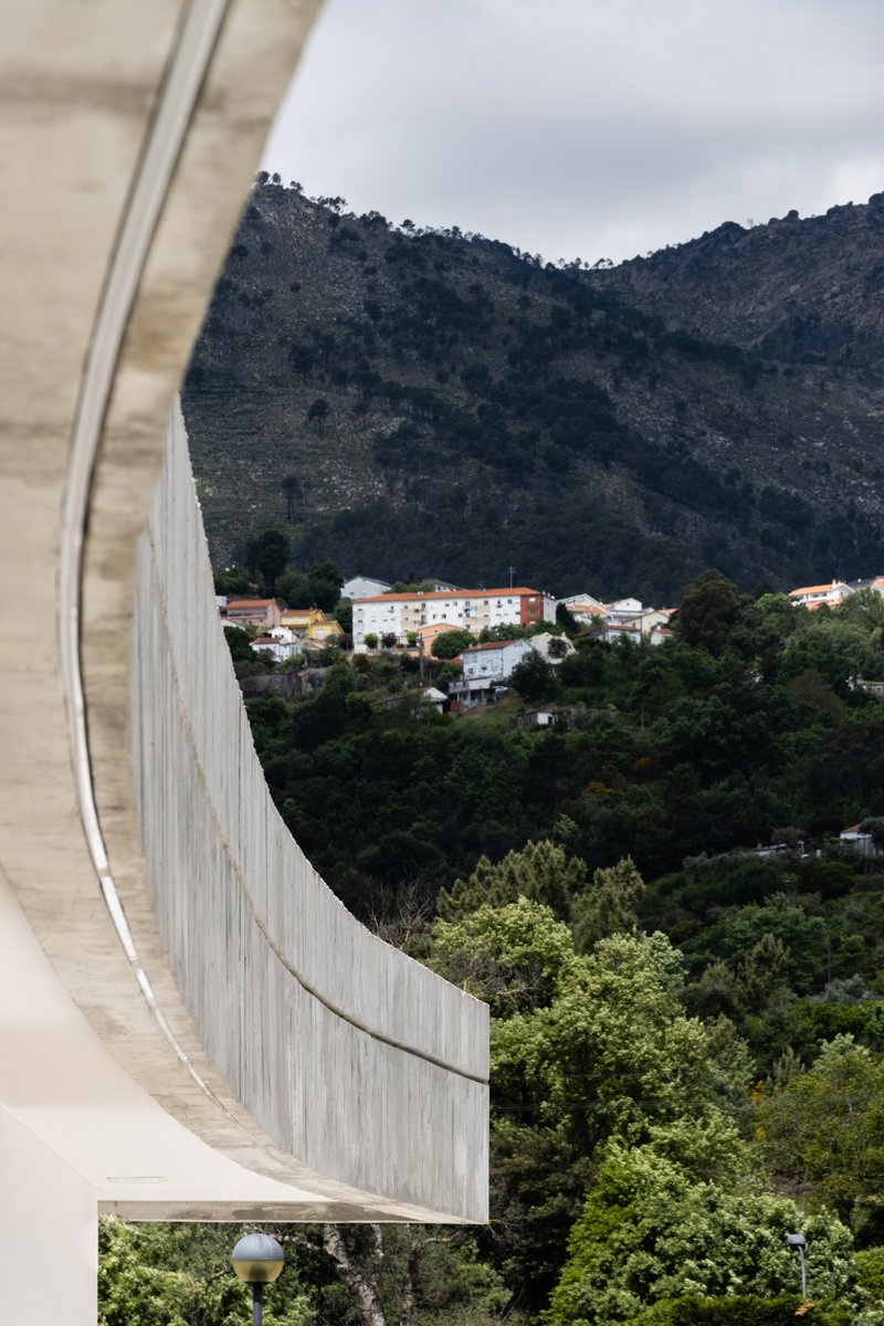 Curved board-formed concrete parapet framing a distant hillside village through green foliage