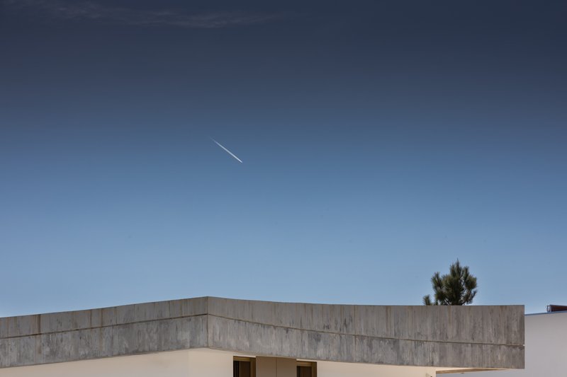 Board-formed concrete parapet with recessed openings below against a clear sky with contrail