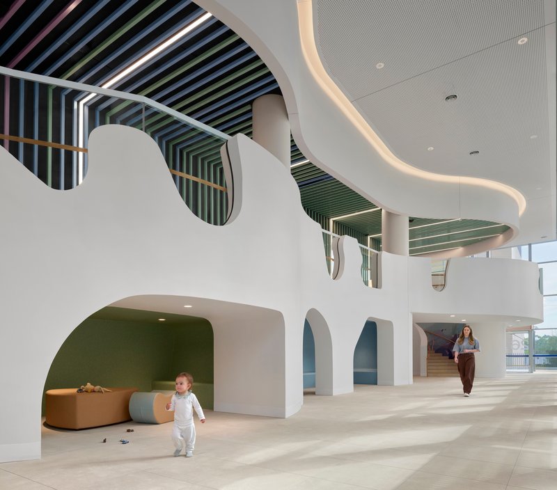 Atrium with curved white walls, arched openings, and rainbow-striped ceiling baffles with child running below
