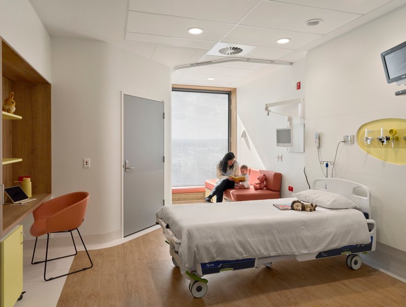 Patient room with hospital bed, window seat, and wooden desk under a suspended ceiling with recessed lighting