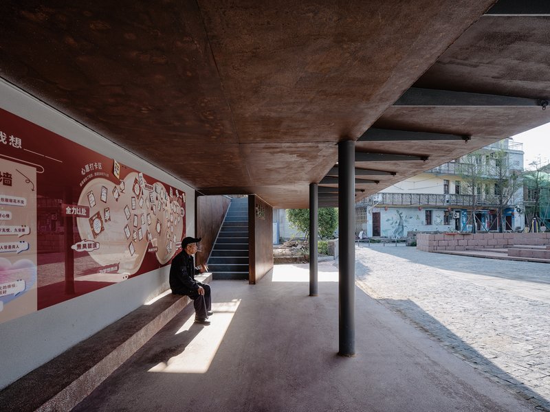 Covered walkway with exposed concrete ceiling, steel columns and a person seated along the red and white mural wall
