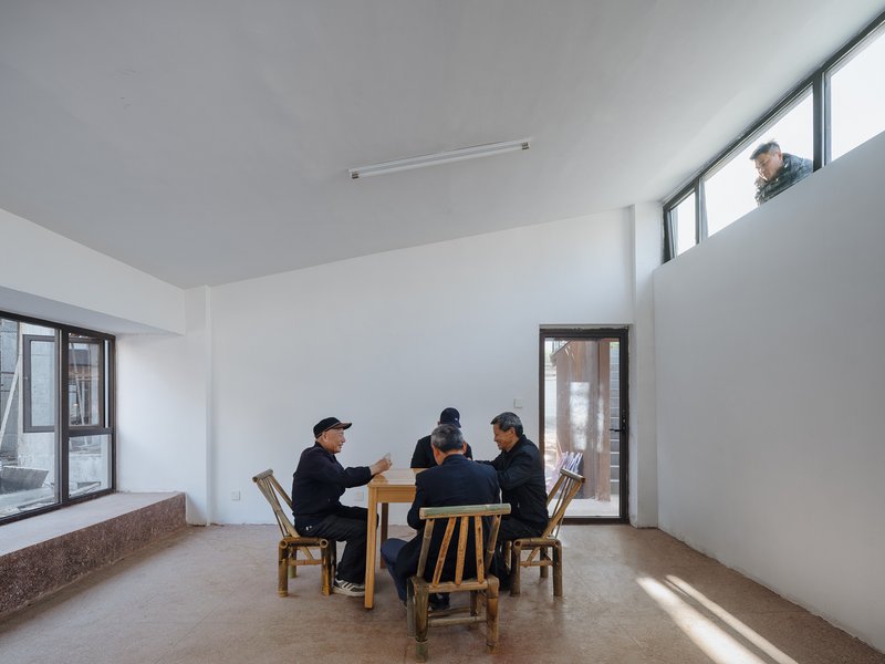 Double-height interior space with people seated at a timber table and a figure visible at the upper window