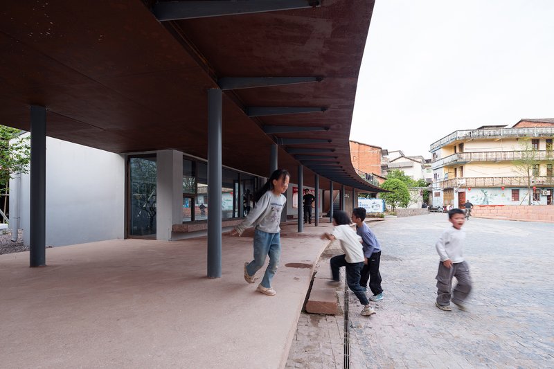 Covered colonnade with timber ceiling panels and children playing in the open plaza beyond the columns