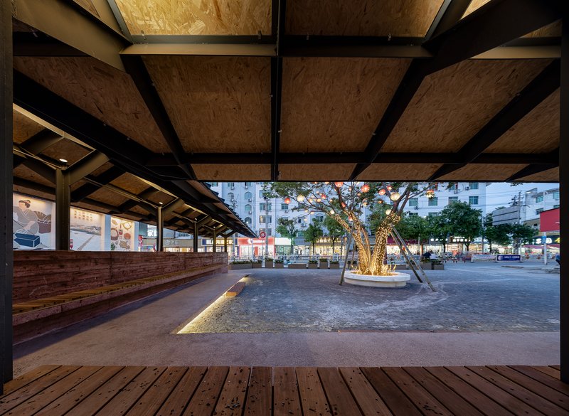 Covered timber deck with plywood ceiling panels and black steel beams overlooking a plaza at twilight