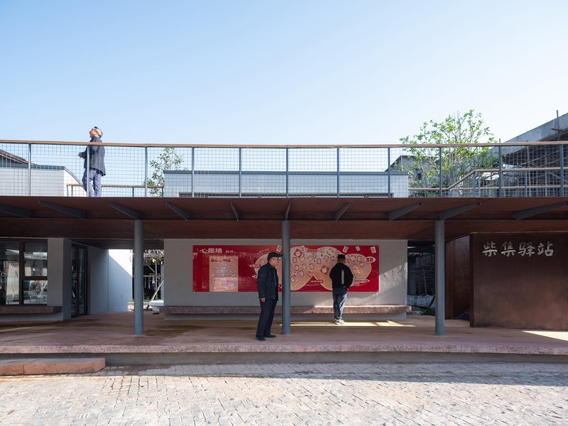 Elevated steel walkway with wire mesh railing above a covered community space at dusk