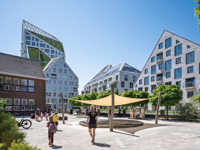 Public plaza with tensile shade canopy and residents walking among the gabled residential towers on a sunny day