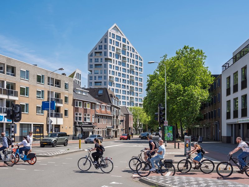Distant view of the gabled tower from a tree-lined street intersection with cyclists crossing in the foreground
