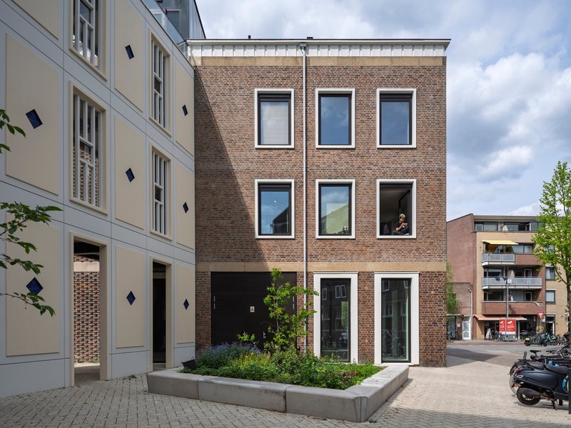 Brown brick facade with symmetrical white-framed windows and planted terrace beside pale neighboring facade