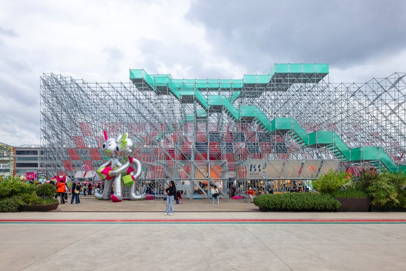 Tubular steel scaffolding structure with turquoise stairs and visitors on the plaza under cloudy skies
