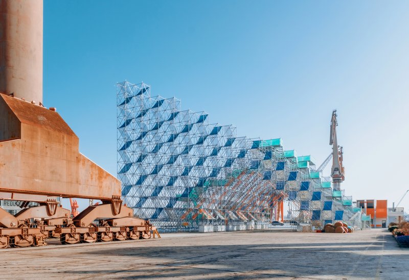 Stepped pyramidal scaffolding structure with blue and green panels beside a wharf and industrial cranes