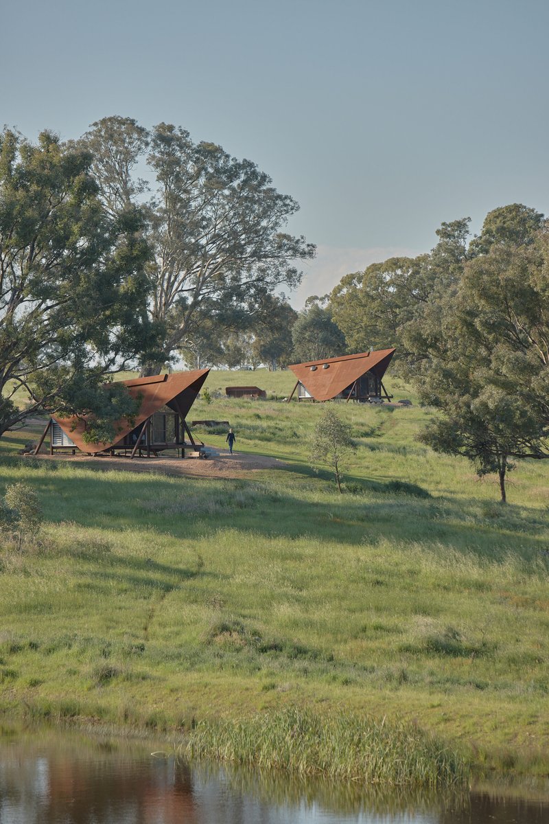Three tented cabins with rust-colored roofs scattered across a sloping grassy field with a pond
