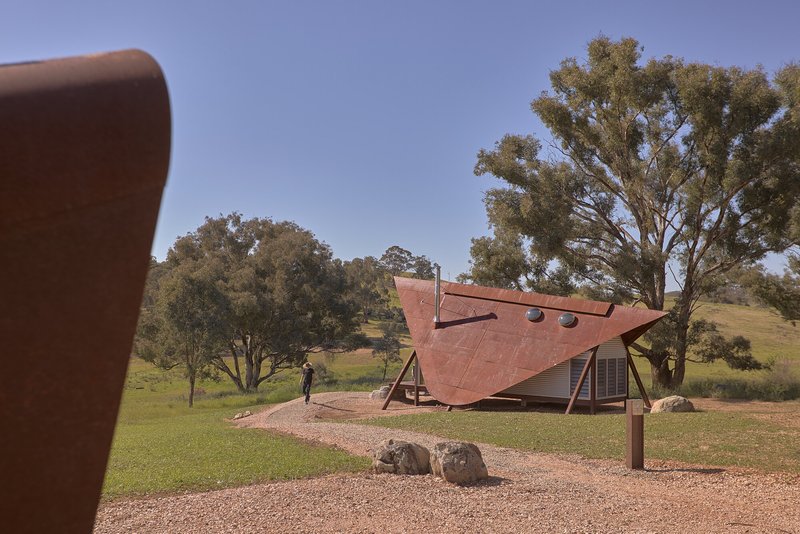 Weathered steel canopy sheltering a glazed cabin wall with a figure walking on gravel beneath