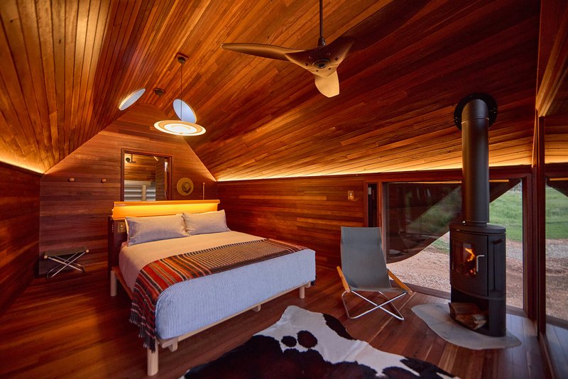 Wood-clad bedroom interior with freestanding stove and cowhide rug at dusk