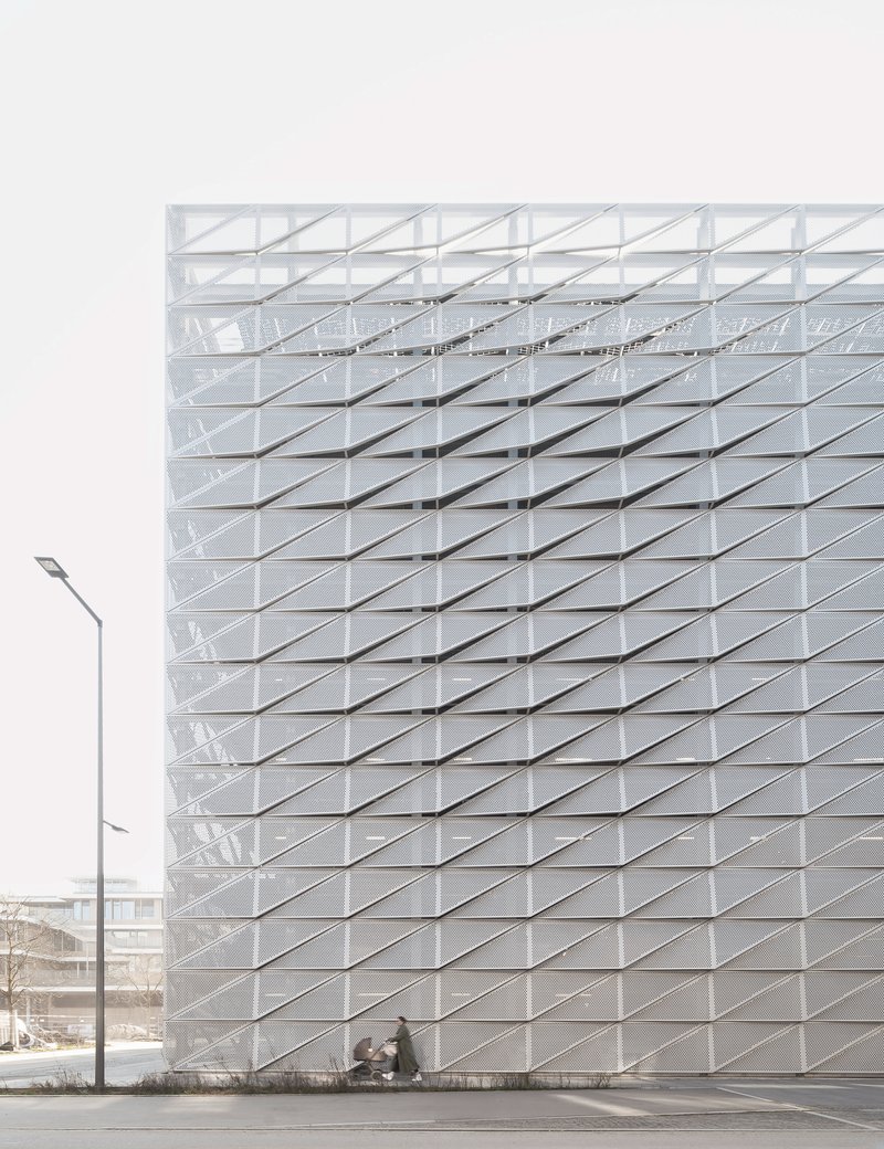 White folded panel facade with diagonal grid pattern and pedestrians at ground level under overcast sky
