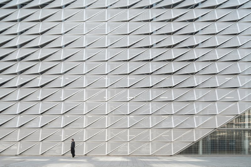 Facade wall of repeating triangular metal panels with a pedestrian standing near the glazed entrance