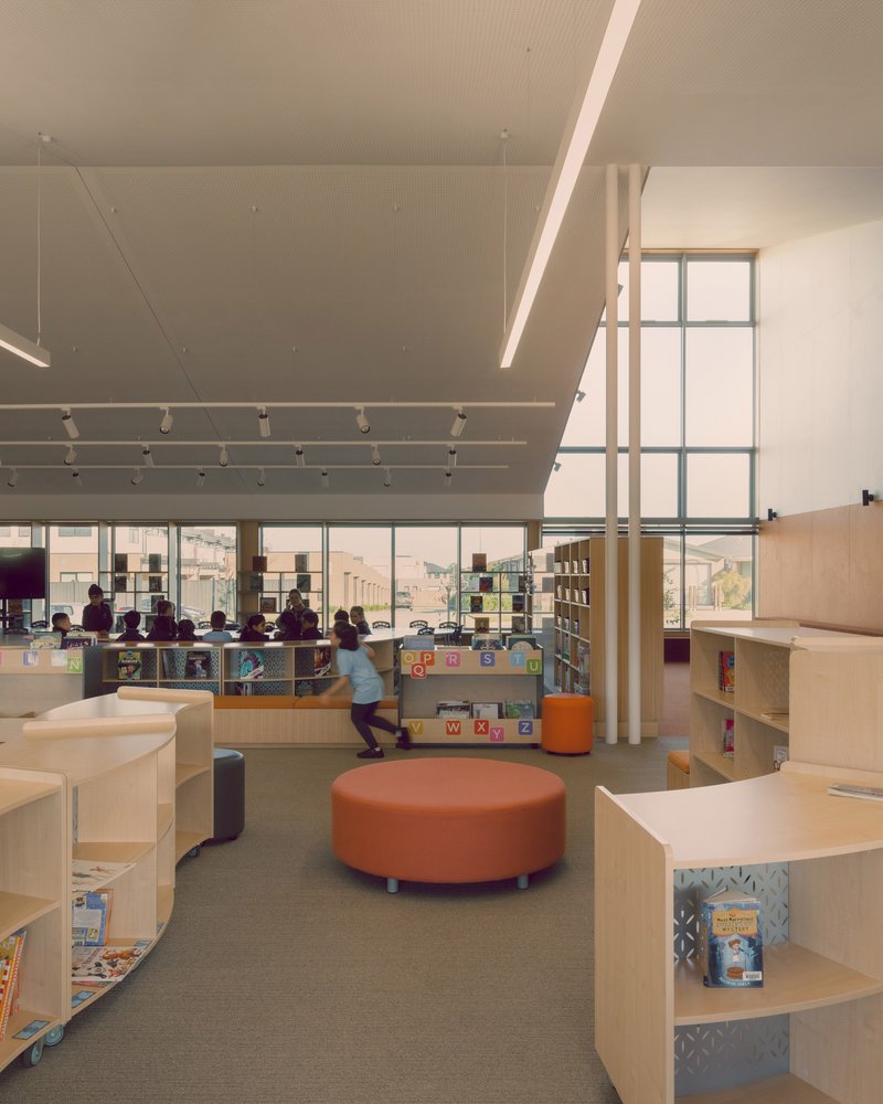 Double-height library interior with perforated ceiling panels, clerestory windows and students browsing low plywood shelving