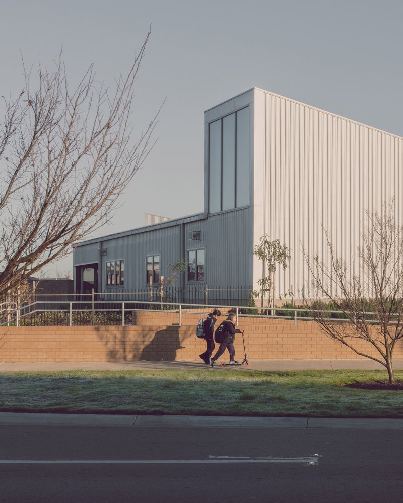 Vertical tower clad in cream corrugated metal rising above horizontal classroom wing with two students walking past