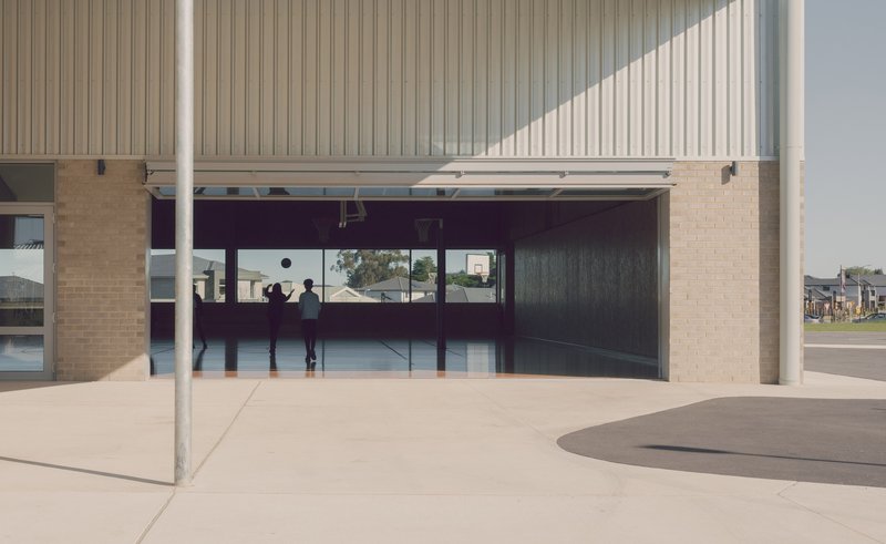 Covered breezeway with cream brick walls, vertical metal cladding and deep eaves framing views to the interior basketball court