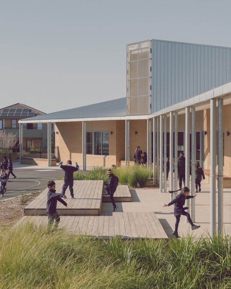 Covered walkway with slender white columns sheltering students playing on timber deck platforms and paving