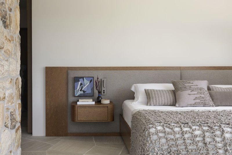 Upholstered headboard with integrated timber and fabric nightstand against a white wall beside rough stone