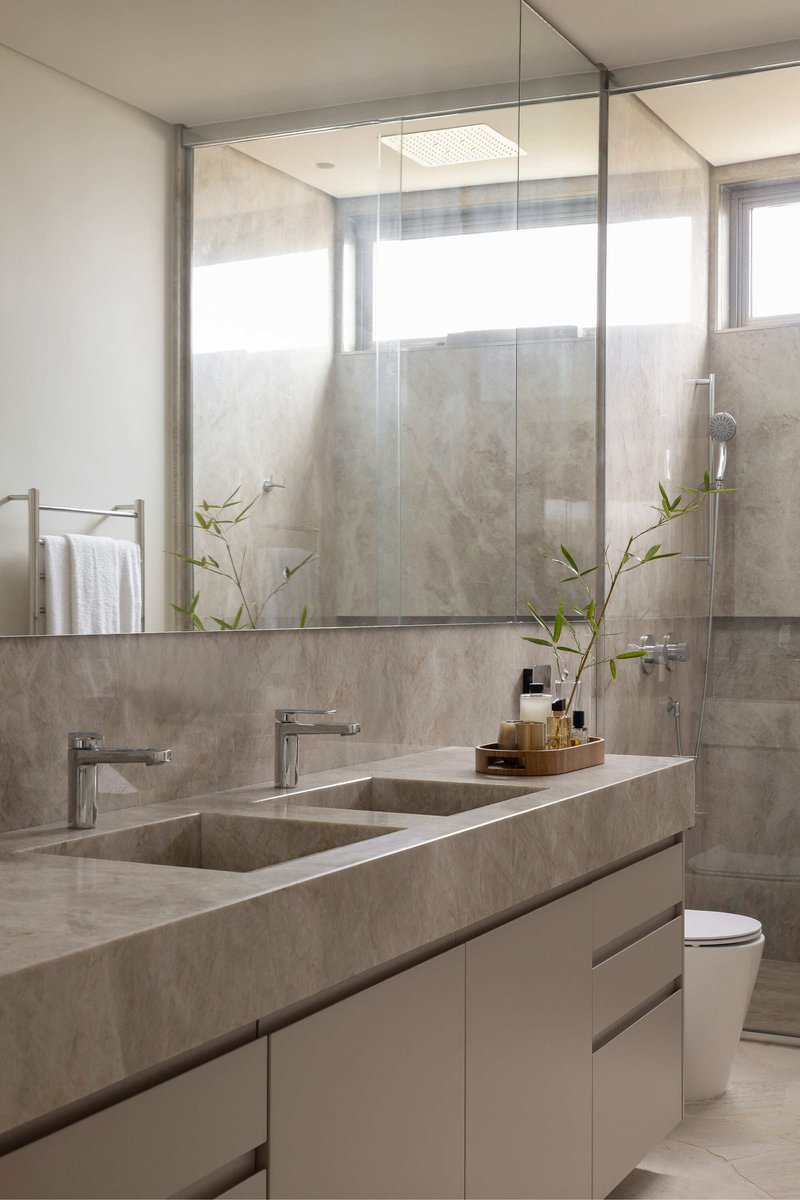 Bathroom vanity with grey stone countertop, twin sinks, and mirror reflecting glass shower enclosure with greenery