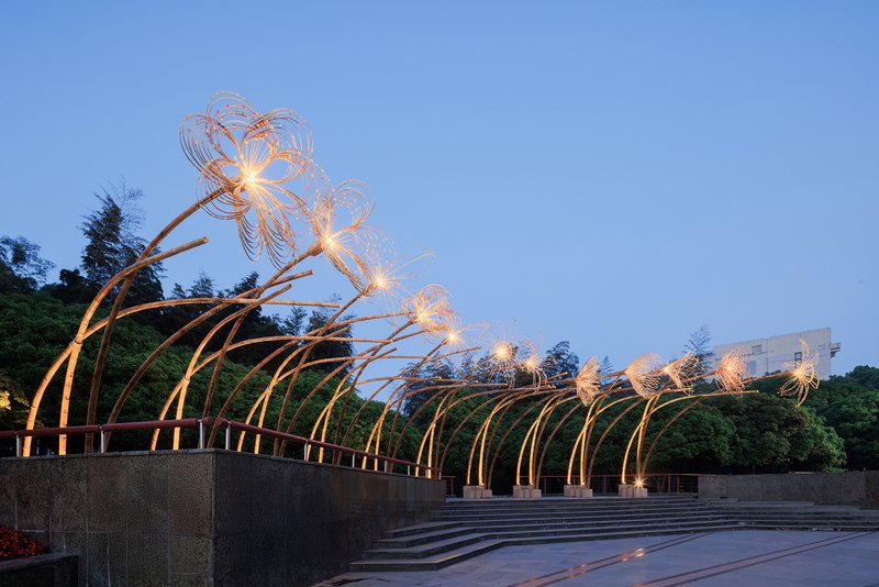 Illuminated steel arch installation with wire blossom forms spanning a public plaza at dusk