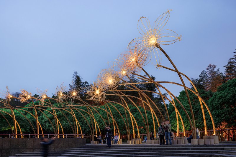 Arched steel structure with lit wire flowers and visitors walking beneath at evening