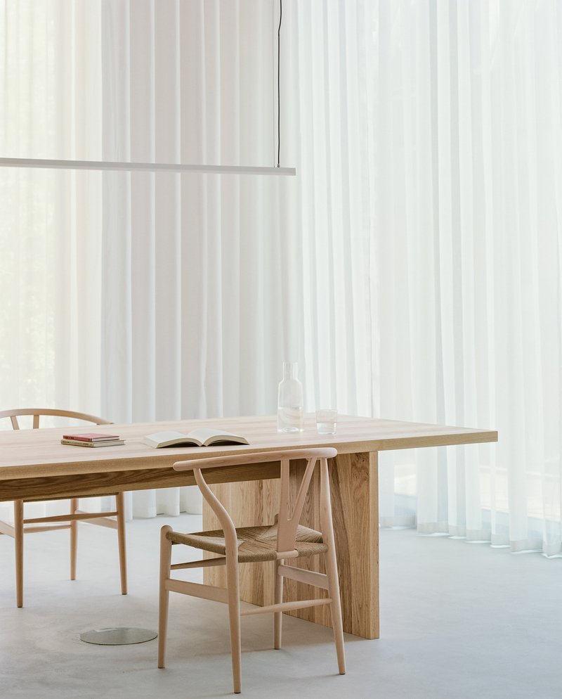 Close view of timber dining table with open book and linear pendant against backlit sheer curtains