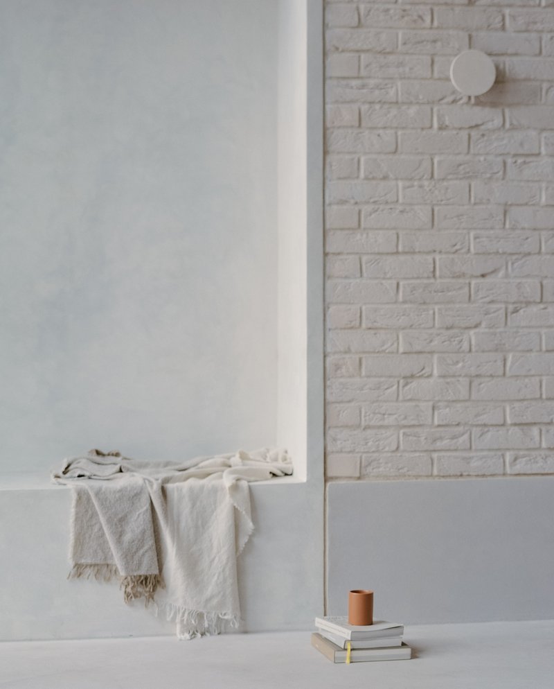 White brick wall with circular sconce above a ledge holding folded linens and stacked books