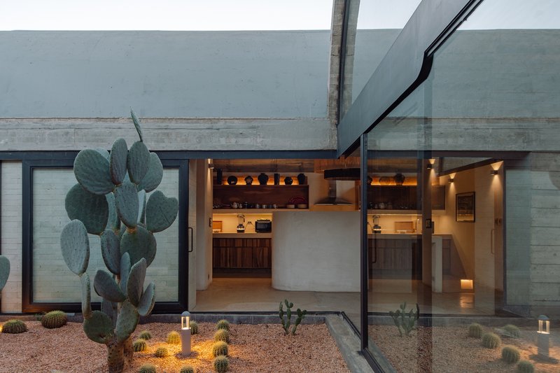 Glass-walled interior kitchen and dining space overlooking a gravel courtyard with cacti illuminated at dusk