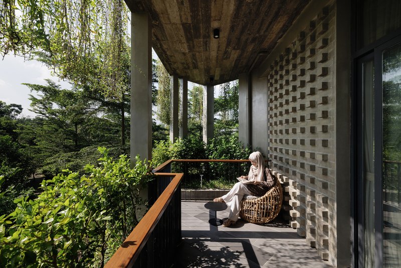 Covered terrace with perforated brick screen, timber railing, and seated figure overlooking treetops in afternoon light