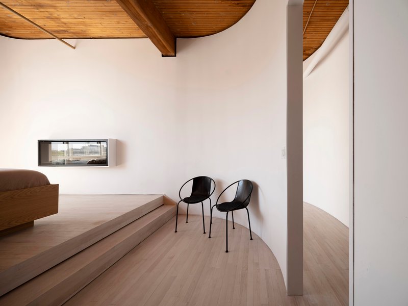 Curved platform with two black chairs beside a low horizontal window under vaulted timber ceiling