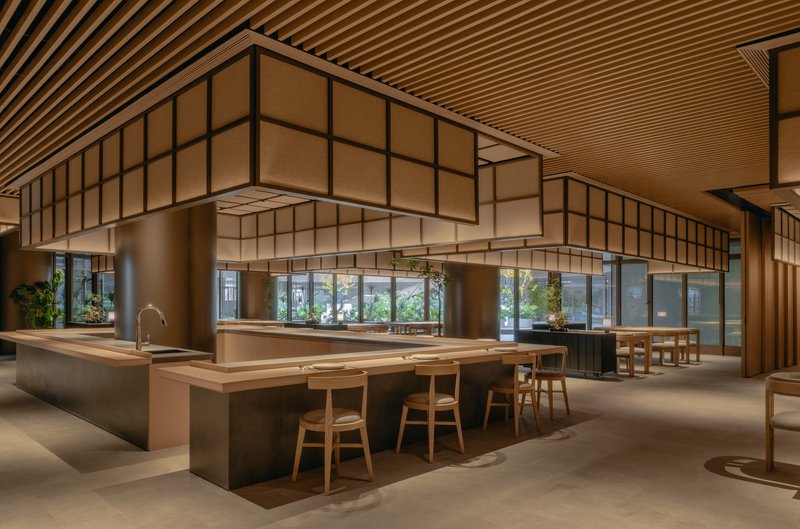 Long interior view showing communal timber table, slatted ceiling and glazed storefront opening to exterior greenery