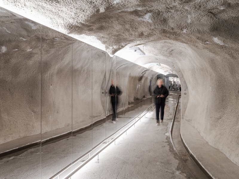 Curved ice tunnel with polished rock walls, glass balustrade, and recessed rail lighting