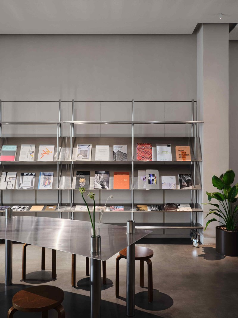Freestanding magazine display unit with publications on angled shelves beside a stainless steel communal table and potted plants